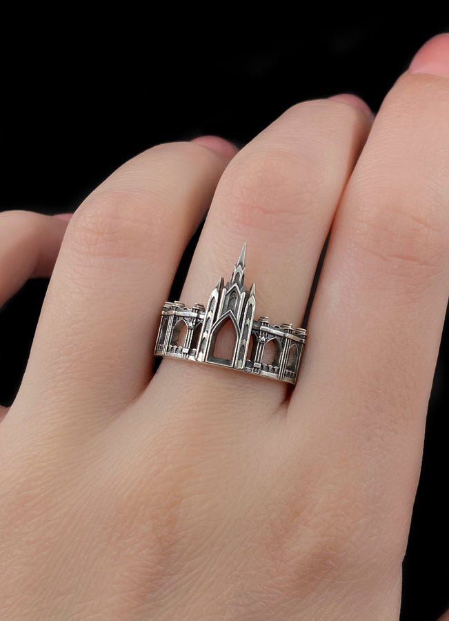 A cinematic studio shot of a sculptural sterling silver ring standing on a reflective surface. The ring is shaped like a miniature building, featuring a tall central spire that rises to a sharp point. The band itself is formed by a colonnade of pointed Gothic arches and buttresses that wrap around the finger. The silver is heavily oxidized, creating deep shadows in the "windows" and "doorways" of the structure. (etsy image id: 7450125356)