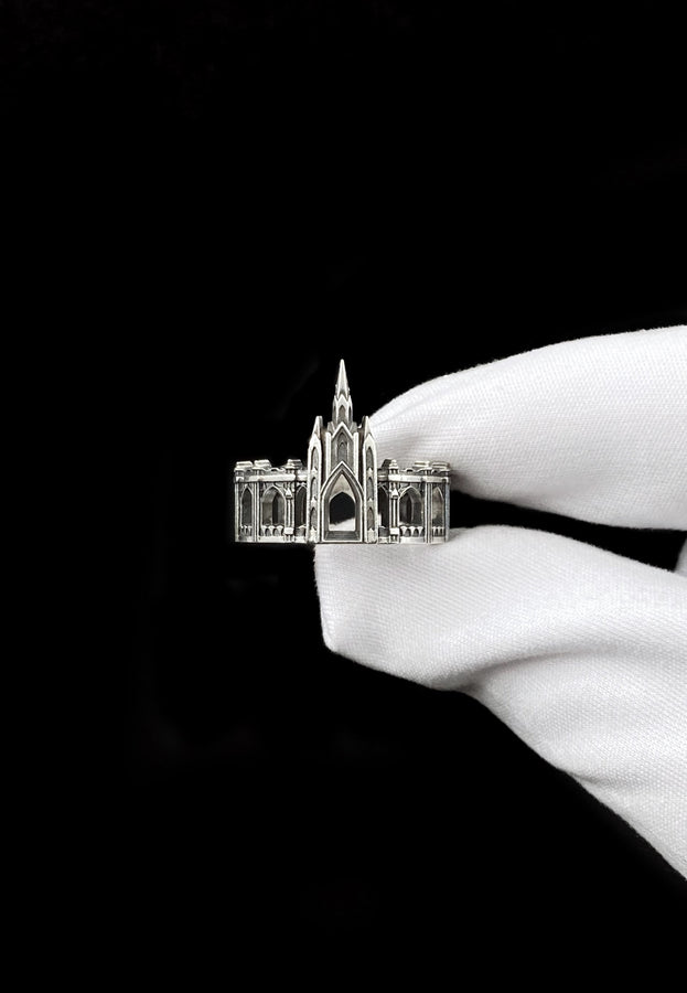A cinematic studio shot of a sculptural sterling silver ring standing on a reflective surface. The ring is shaped like a miniature building, featuring a tall central spire that rises to a sharp point. The band itself is formed by a colonnade of pointed Gothic arches and buttresses that wrap around the finger. The silver is heavily oxidized, creating deep shadows in the "windows" and "doorways" of the structure. (etsy image id: 7450125356)