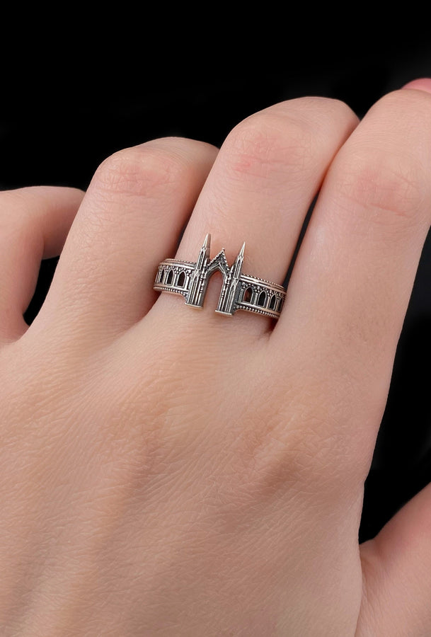 A cinematic studio shot of a sculptural sterling silver ring standing on a reflective surface. The design mimics the facade of a Gothic cathedral or castle gate. Two tall, pointed spires rise up on either side of a central archway. The band continues outwards with a colonnade of smaller arches. The silver features a beaded "cornice" detail along the edges and is heavily oxidized to emphasize the depth of the "stonework." (etsy image id: 7450300962)