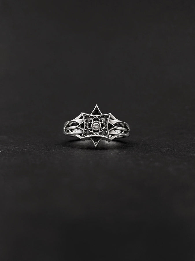 A detailed studio shot of a sterling silver ring held in a gloved hand. The focal point is a geometric "North Star" face—a square frame turned on its point, embellished with top and bottom spikes. Inside the frame is a floral quatrefoil pattern surrounding a round, bezel-set white gemstone (Diamond or CZ). The silver is polished on the raised lines and dark in the recesses. (etsy image id: 7452221522)
