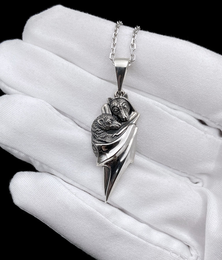 A detailed close-up studio shot of a sterling silver pendant shaped like two flying foxes or fruit bats. The bats are intimately huddled together, wrapped within the smooth, sweeping folds of their leathery wings. The metal is highly textured to show the fine details of their fur and faces. The silver is heavily oxidized in the recesses, creating a striking contrast against the polished highlights of the wings. (etsy image id: 5592925539)