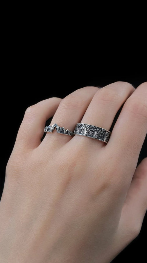 A cinematic studio shot of two matching sterling silver rings. The top ring is a delicate "crown" or tiara shape, formed by the peak of a Gothic arch with trefoil tracery. The bottom ring is a wide band featuring a continuous row of identical arches. The background metal inside the arches has a rough, "eroded" or stippled texture, heavily oxidized to black, while the raised architectural lines are polished smooth.