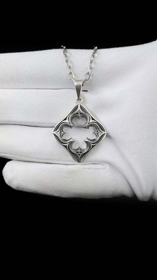 A cinematic studio shot of a diamond-shaped sterling silver pendant hanging against a smoky, amber-lit background. The design is "openwork," featuring a central cutout in the shape of a four-lobed quatrefoil. The silver frame mimics the stone mullions of a Gothic window, with pointed arches and sharp cusps. The metal is polished on the ridges and darkened on the sides to define the shape against the void. (etsy image id: 7496323581)