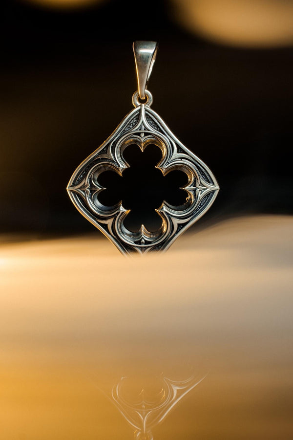 A cinematic studio shot of a diamond-shaped sterling silver pendant hanging against a smoky, amber-lit background. The design is "openwork," featuring a central cutout in the shape of a four-lobed quatrefoil. The silver frame mimics the stone mullions of a Gothic window, with pointed arches and sharp cusps. The metal is polished on the ridges and darkened on the sides to define the shape against the void. (etsy image id: 7496323581)