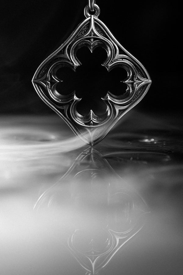 A cinematic studio shot of a diamond-shaped sterling silver pendant hanging against a smoky, amber-lit background. The design is "openwork," featuring a central cutout in the shape of a four-lobed quatrefoil. The silver frame mimics the stone mullions of a Gothic window, with pointed arches and sharp cusps. The metal is polished on the ridges and darkened on the sides to define the shape against the void. (etsy image id: 7496323581)