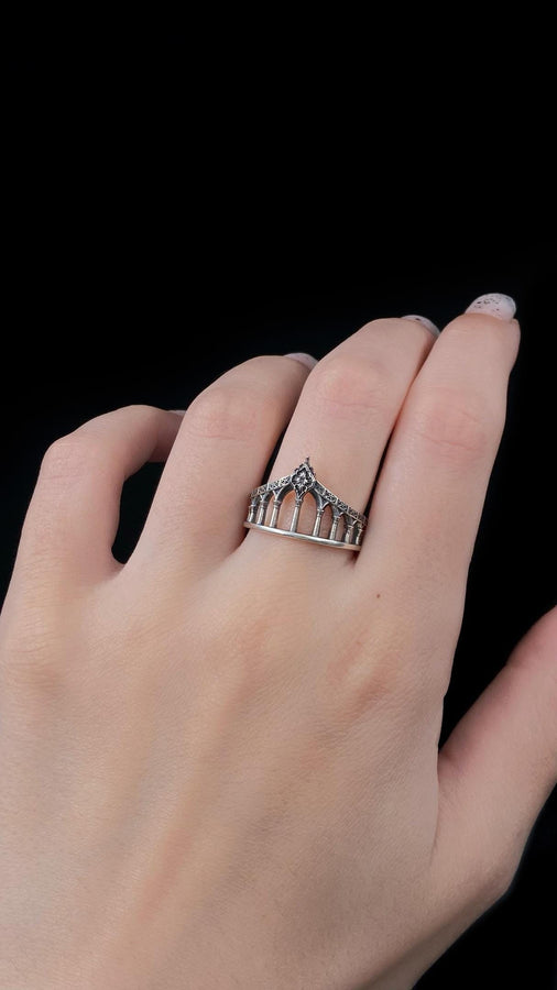 A cinematic studio shot of a sculptural sterling silver ring standing in shallow water. The design mimics a bridge or aqueduct, featuring a row of vertical pillars or columns that support a rising upper rail. The center rises to a sharp point adorned with a floral or quatrefoil relief. The space between the columns is open, creating a "see-through" architectural silhouette. (etsy image id: 7450247674)