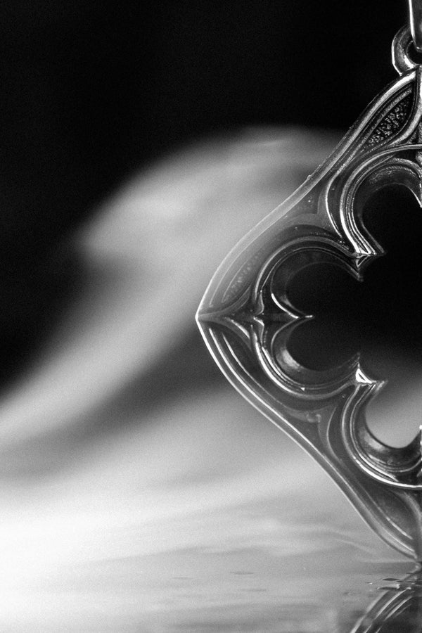 A cinematic studio shot of a diamond-shaped sterling silver pendant hanging against a smoky, amber-lit background. The design is "openwork," featuring a central cutout in the shape of a four-lobed quatrefoil. The silver frame mimics the stone mullions of a Gothic window, with pointed arches and sharp cusps. The metal is polished on the ridges and darkened on the sides to define the shape against the void. (etsy image id: 7496323581)