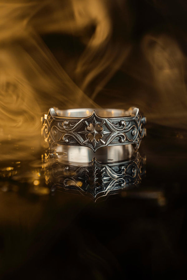 A cinematic studio shot of a matching two-piece ring set on dark velvet. The top ring stands upright, featuring a diamond-shaped face with a four-pointed star design and a small, round black stone in the center. The bottom ring lies flat, displaying a wide band with a repeating pattern of the same star/cross motif connected by swirling filigree. The silver is polished on the ridges and dark in the background.