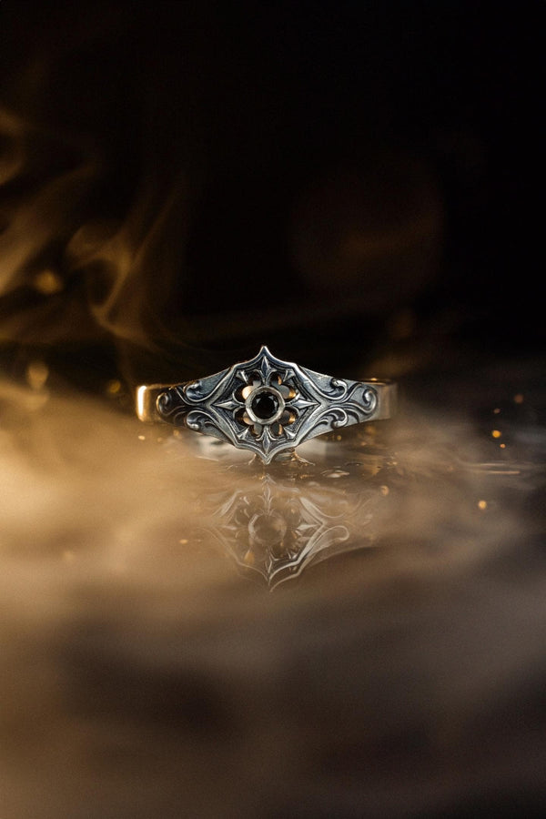 A cinematic studio shot of a matching two-piece ring set on dark velvet. The top ring stands upright, featuring a diamond-shaped face with a four-pointed star design and a small, round black stone in the center. The bottom ring lies flat, displaying a wide band with a repeating pattern of the same star/cross motif connected by swirling filigree. The silver is polished on the ridges and dark in the background.