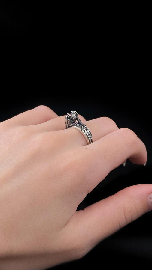 A detailed profile shot of a sculptural sterling silver ring. The band is formed by the body of a crouching gargoyle. The creature features distinct horns, a snarling face, and muscular limbs. Its bat-like wings fold back to form the sides of the ring shank. In its front claw, it holds a small, circular charm with a web pattern, resembling a dreamcatcher. The silver is heavily oxidized black in the crevices to accentuate the details. (etsy image id: 7497960785)