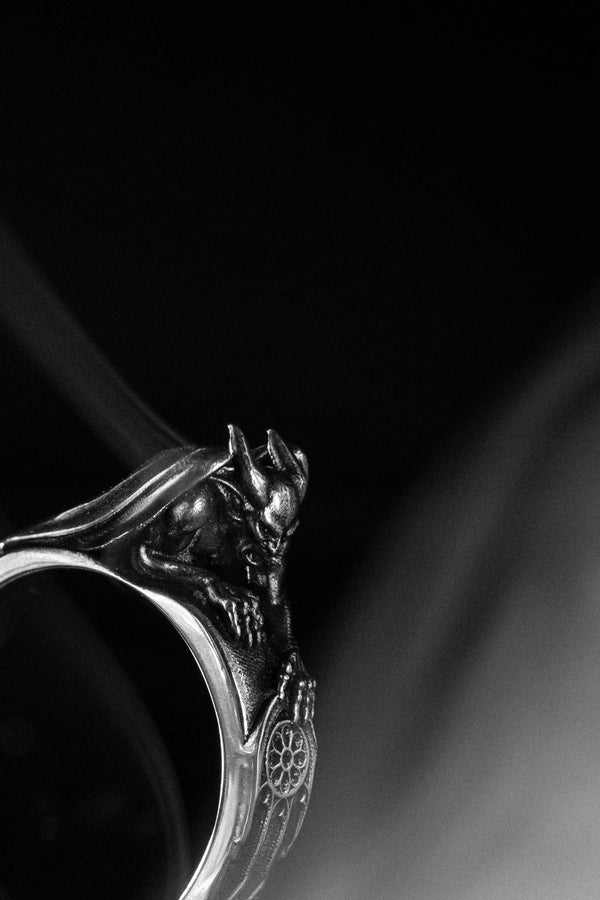 A detailed profile shot of a sculptural sterling silver ring. The band is formed by the body of a crouching gargoyle. The creature features distinct horns, a snarling face, and muscular limbs. Its bat-like wings fold back to form the sides of the ring shank. In its front claw, it holds a small, circular charm with a web pattern, resembling a dreamcatcher. The silver is heavily oxidized black in the crevices to accentuate the details. (etsy image id: 7497960785)