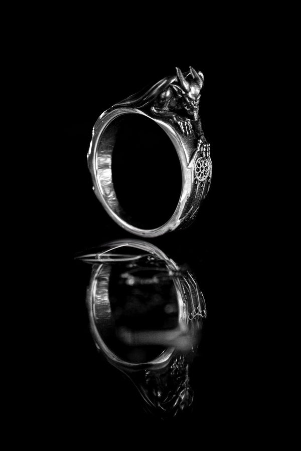 A detailed profile shot of a sculptural sterling silver ring. The band is formed by the body of a crouching gargoyle. The creature features distinct horns, a snarling face, and muscular limbs. Its bat-like wings fold back to form the sides of the ring shank. In its front claw, it holds a small, circular charm with a web pattern, resembling a dreamcatcher. The silver is heavily oxidized black in the crevices to accentuate the details. (etsy image id: 7497960785)
