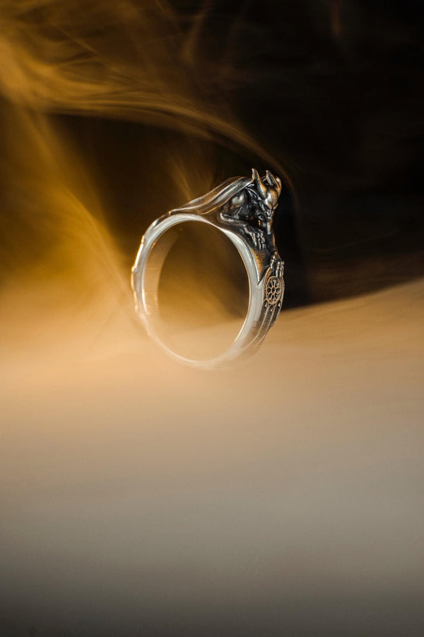 A detailed profile shot of a sculptural sterling silver ring. The band is formed by the body of a crouching gargoyle. The creature features distinct horns, a snarling face, and muscular limbs. Its bat-like wings fold back to form the sides of the ring shank. In its front claw, it holds a small, circular charm with a web pattern, resembling a dreamcatcher. The silver is heavily oxidized black in the crevices to accentuate the details. (etsy image id: 7497960785)