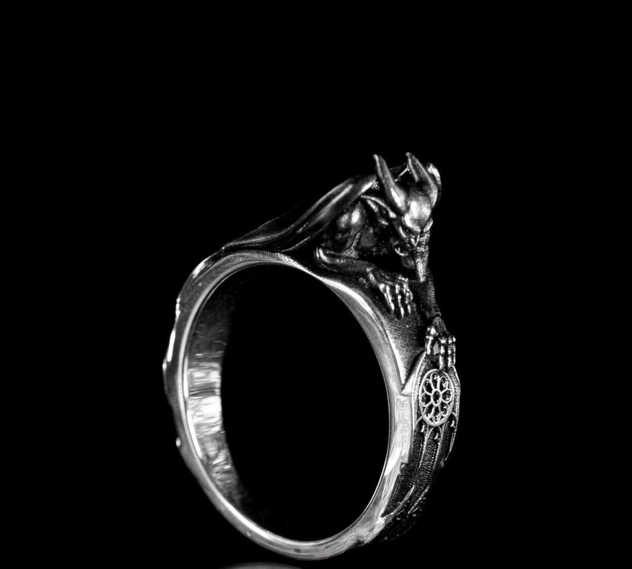 A detailed profile shot of a sculptural sterling silver ring. The band is formed by the body of a crouching gargoyle. The creature features distinct horns, a snarling face, and muscular limbs. Its bat-like wings fold back to form the sides of the ring shank. In its front claw, it holds a small, circular charm with a web pattern, resembling a dreamcatcher. The silver is heavily oxidized black in the crevices to accentuate the details. (etsy image id: 7497960785)