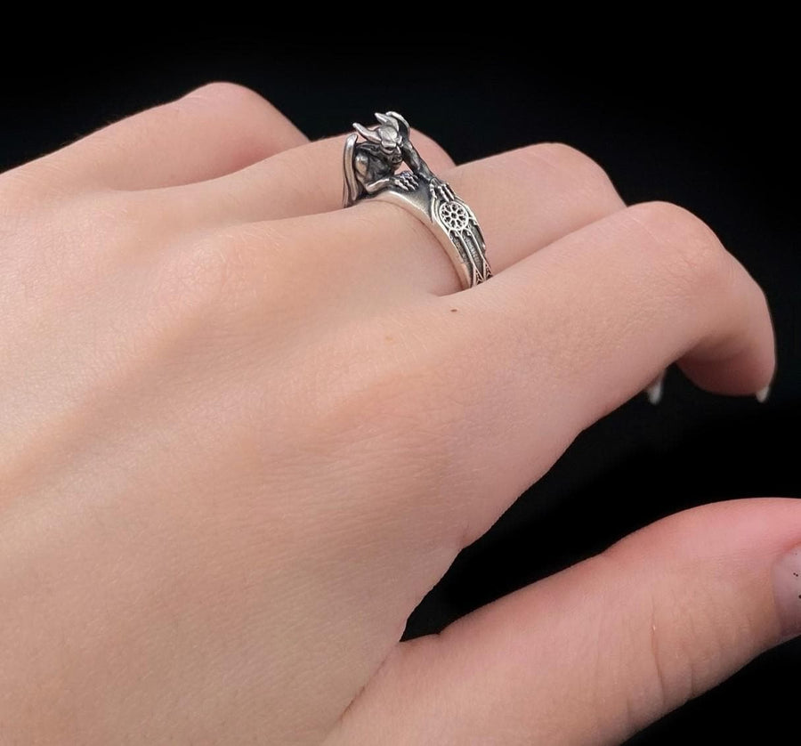 A detailed profile shot of a sculptural sterling silver ring. The band is formed by the body of a crouching gargoyle. The creature features distinct horns, a snarling face, and muscular limbs. Its bat-like wings fold back to form the sides of the ring shank. In its front claw, it holds a small, circular charm with a web pattern, resembling a dreamcatcher. The silver is heavily oxidized black in the crevices to accentuate the details. (etsy image id: 7497960785)