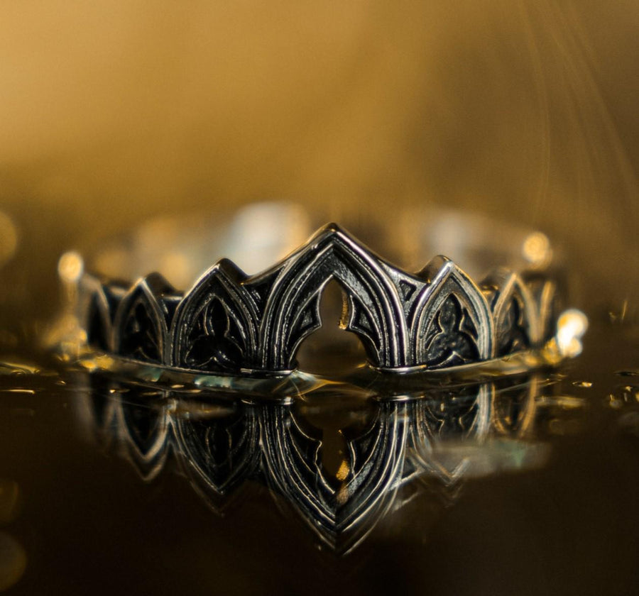 A cinematic studio shot of two matching sterling silver rings. The top ring is a delicate "crown" or tiara shape, formed by the peak of a Gothic arch with trefoil tracery. The bottom ring is a wide band featuring a continuous row of identical arches. The background metal inside the arches has a rough, "eroded" or stippled texture, heavily oxidized to black, while the raised architectural lines are polished smooth.