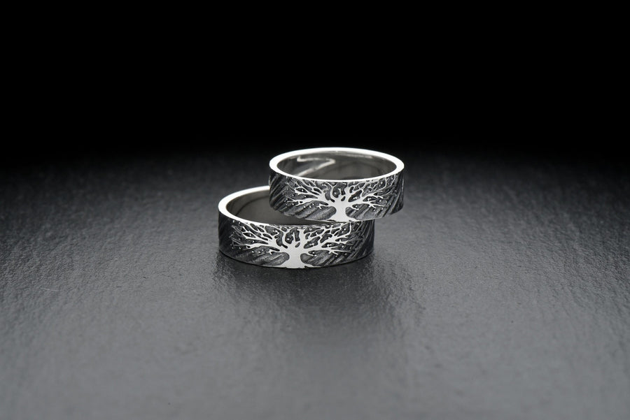 A clean studio shot of two matching sterling silver rings resting on a slate grey surface. Both bands feature a continuous panoramic design of a tree with spreading branches and deep roots. The tree motif is polished silver, which contrasts sharply with the textured, oxidized grey background that resembles tree bark or drifting soil.