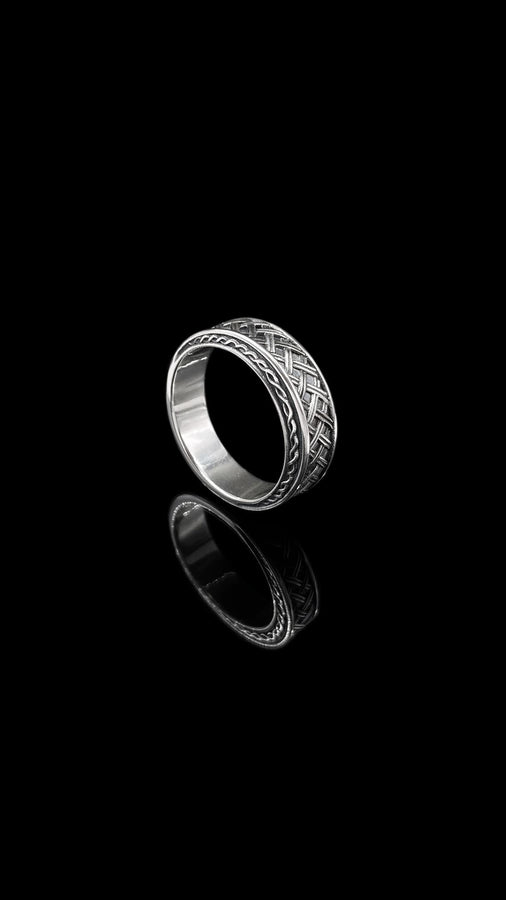 A warm, atmospheric shot of a matching wedding ring set. The engagement ring stands upright, showcasing a round white gemstone in a bezel setting. Lying flat beneath it is a matching wide band without a stone. Both rings share a cohesive design: a central diagonal "basket weave" pattern flanked by twisted rope borders. The silver is oxidized to reveal the intricate texture of the weave.
