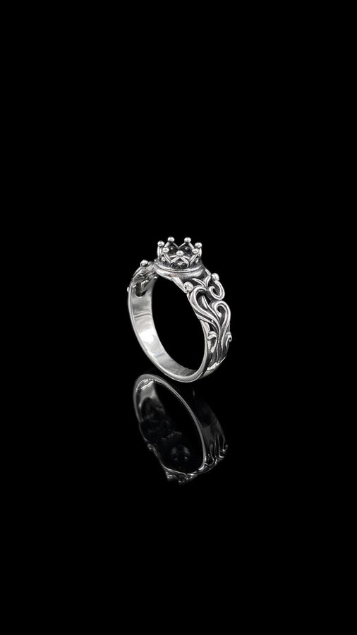 A high-contrast studio shot of a two-piece sterling silver ring set. The top ring features a raised, three-dimensional "crown" or "coronet" motif with small spheres on the tips. The bottom ring is a matching wide band. Both rings are covered in intricate, raised scrollwork or vine-like filigree. The silver is polished bright on the raised swirls, contrasting with the black oxidized background.