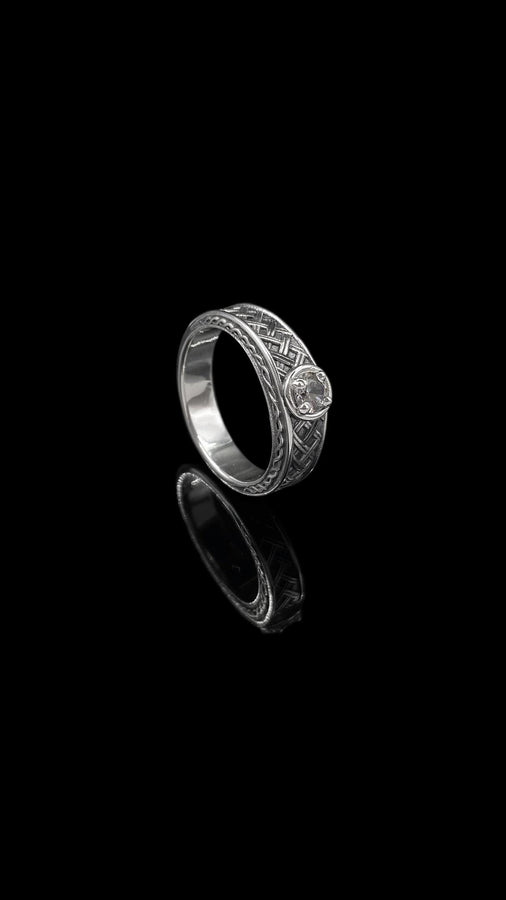 A warm, atmospheric shot of a matching wedding ring set. The engagement ring stands upright, showcasing a round white gemstone in a bezel setting. Lying flat beneath it is a matching wide band without a stone. Both rings share a cohesive design: a central diagonal "basket weave" pattern flanked by twisted rope borders. The silver is oxidized to reveal the intricate texture of the weave.