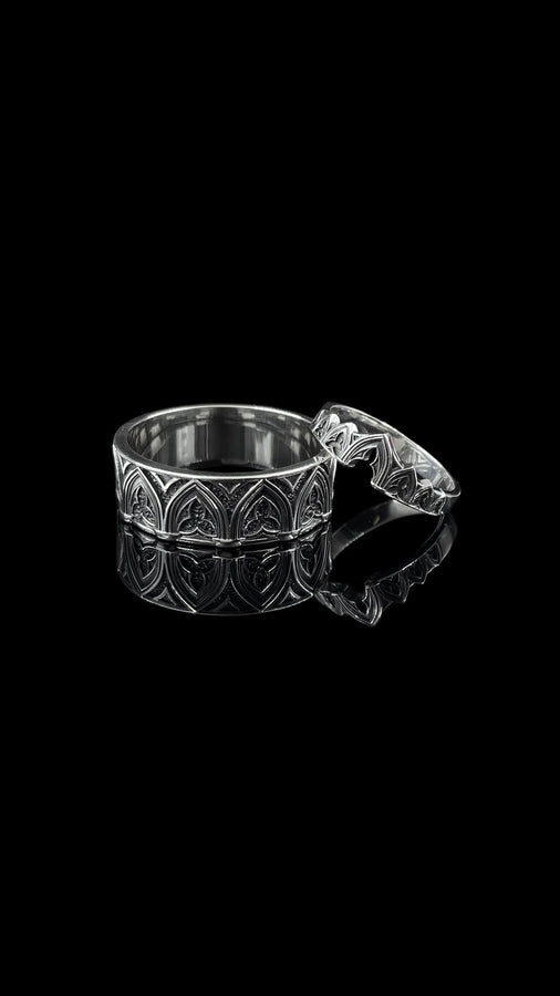 A cinematic studio shot of two matching sterling silver rings. The top ring is a delicate "crown" or tiara shape, formed by the peak of a Gothic arch with trefoil tracery. The bottom ring is a wide band featuring a continuous row of identical arches. The background metal inside the arches has a rough, "eroded" or stippled texture, heavily oxidized to black, while the raised architectural lines are polished smooth.