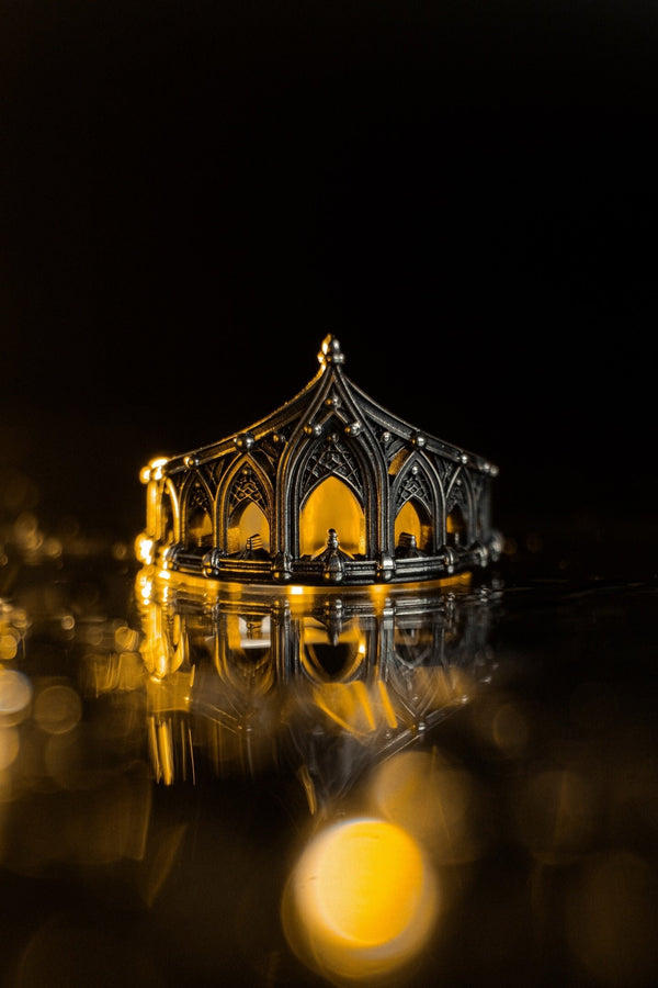 A cinematic studio shot of a sculptural sterling silver ring standing on a reflective surface. The ring is designed as a "crown" or "coronet," formed by a continuous circle of Gothic cathedral arches. The central arch is the tallest, creating a focal point like a spire. The silver is heavily oxidized, with black recesses emphasizing the ribbed vaulting and architectural details against the polished ridges.