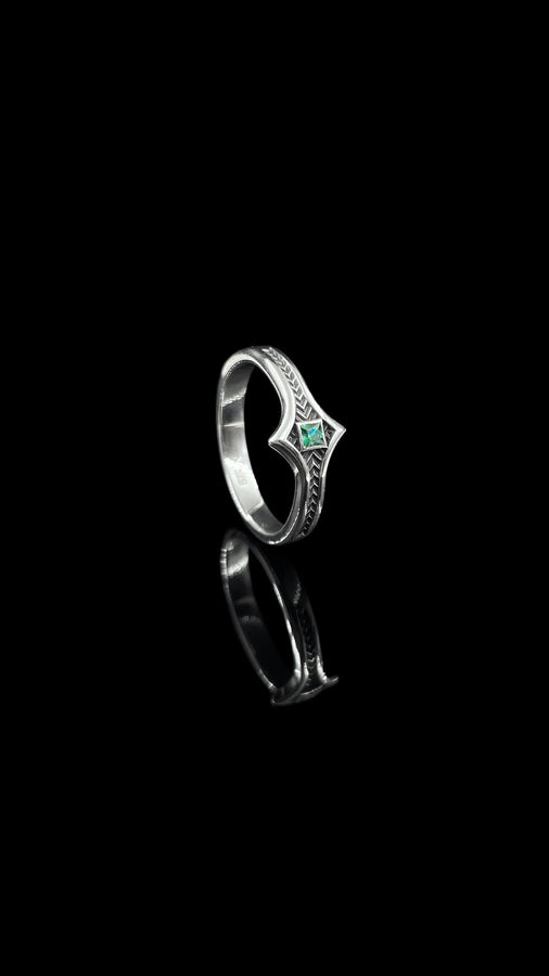 A high-definition shot of a matching two-piece wedding ring set resting on dark velvet. The foreground features a delicate sterling silver ring with a pointed, kite-shaped face holding a square blue gemstone. Standing behind it is a wide, sturdy band with a repeating quatrefoil "clover" pattern. Both rings share a cohesive finish: bright polished silver highlights against a deeply oxidized, matte black background.