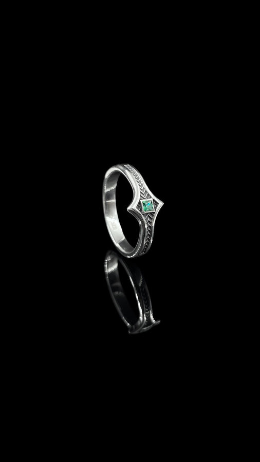 A high-definition shot of a matching two-piece wedding ring set resting on dark velvet. The foreground features a delicate sterling silver ring with a pointed, kite-shaped face holding a square blue gemstone. Standing behind it is a wide, sturdy band with a repeating quatrefoil "clover" pattern. Both rings share a cohesive finish: bright polished silver highlights against a deeply oxidized, matte black background.
