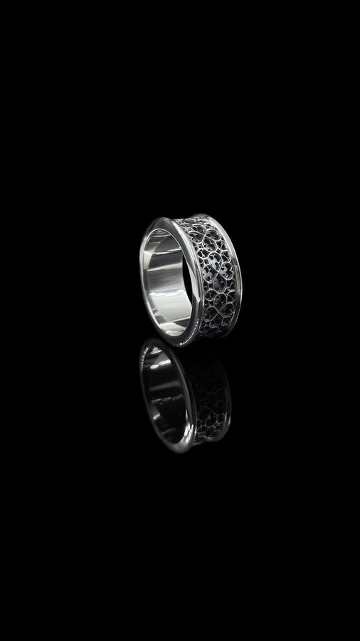 A high-definition shot of a matching two-piece wedding ring set resting on dark velvet. The foreground features a delicate sterling silver ring with a pointed, kite-shaped face holding a square blue gemstone. Standing behind it is a wide, sturdy band with a repeating quatrefoil "clover" pattern. Both rings share a cohesive finish: bright polished silver highlights against a deeply oxidized, matte black background.