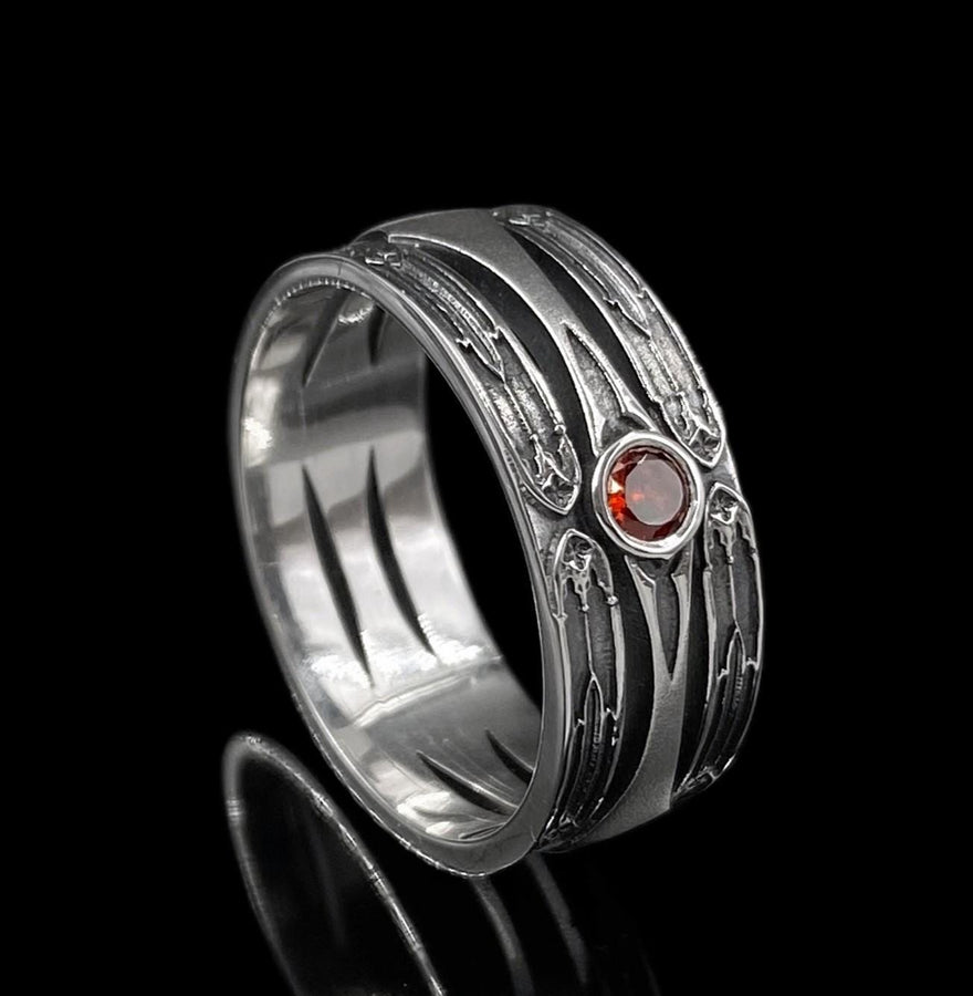 A high-contrast studio shot of a two-piece sterling silver ring set. The foreground features a wide band lying flat, set with a round clear gemstone. Flanking the stone are horizontal, grooved lines resembling arrows or spears. Standing behind it is a narrower engagement-style ring with a matching "lance-tip" shank leading to a central bezel-set white stone. The background is a textured, dark fabric.