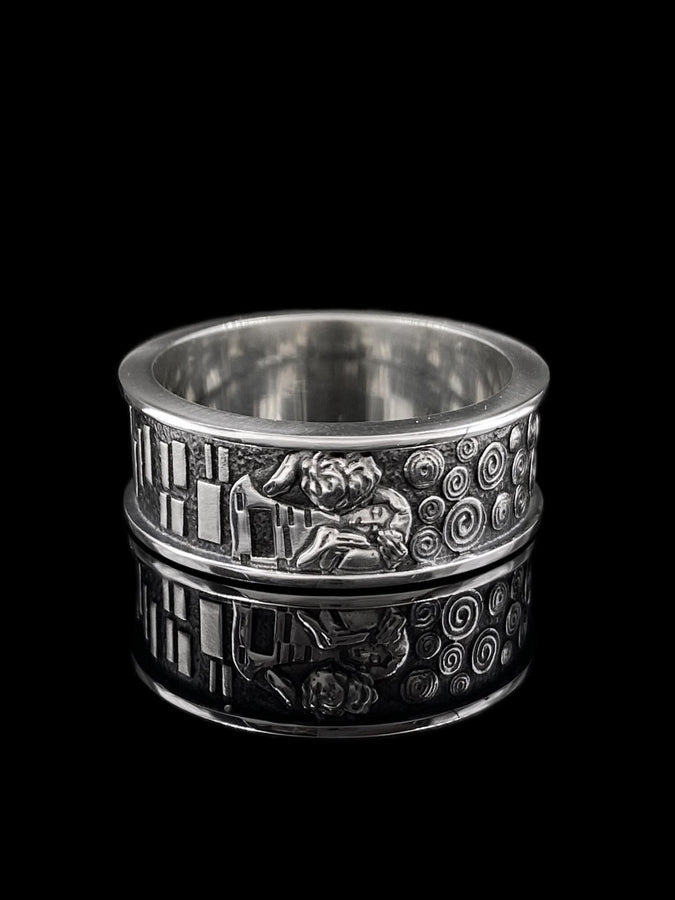 A cinematic studio shot of a matching two-piece ring set. The bottom ring is a wide silver band featuring a detailed relief carving of the couple from Gustav Klimt's "The Kiss." The top ring is a narrower engagement band that mimics the geometric patterns of the painting and holds a round, amber-colored gemstone. The background is a warm, golden haze, reflecting the "Golden Phase" of the original artwork.