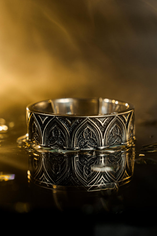 A cinematic studio shot of two matching sterling silver rings. The top ring is a delicate "crown" or tiara shape, formed by the peak of a Gothic arch with trefoil tracery. The bottom ring is a wide band featuring a continuous row of identical arches. The background metal inside the arches has a rough, "eroded" or stippled texture, heavily oxidized to black, while the raised architectural lines are polished smooth.