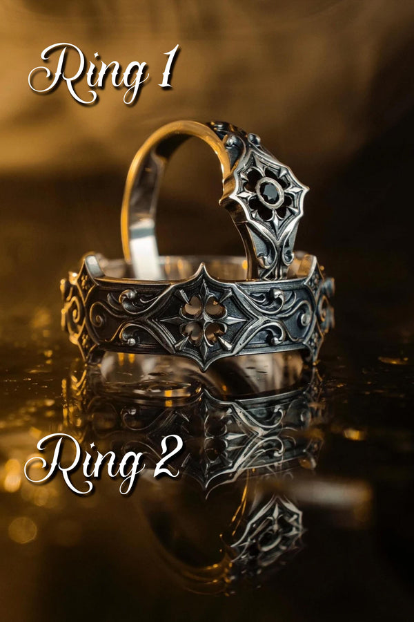 A cinematic studio shot of a matching two-piece ring set on dark velvet. The top ring stands upright, featuring a diamond-shaped face with a four-pointed star design and a small, round black stone in the center. The bottom ring lies flat, displaying a wide band with a repeating pattern of the same star/cross motif connected by swirling filigree. The silver is polished on the ridges and dark in the background.