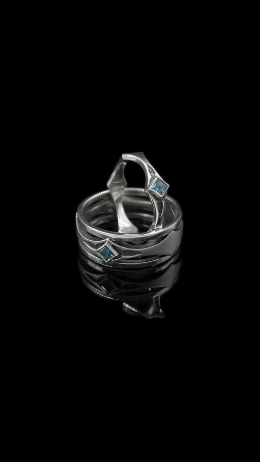 This Art Deco wedding ring set features a sterling silver engagement ring and matching band with bold geometric cutouts and a bright blue square gemstone. The clean lines, layered shapes, and sculptural silhouette create a modern yet vintage feel ideal for couples seeking unique, architecturally inspired bridal jewelry captured on a reflective surface for elegant presentation.