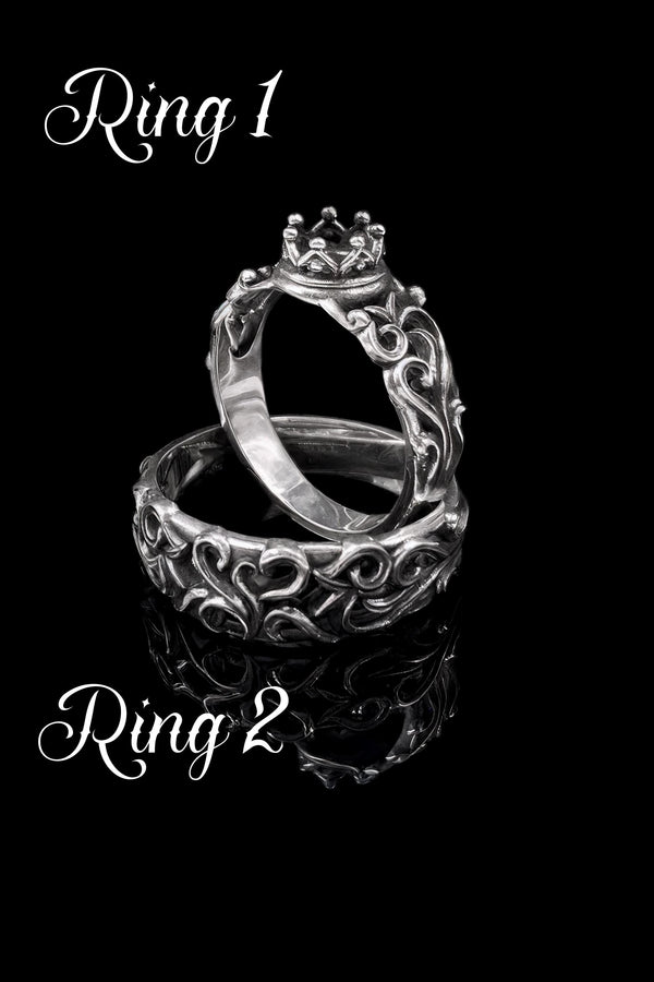 A high-contrast studio shot of a two-piece sterling silver ring set. The top ring features a raised, three-dimensional "crown" or "coronet" motif with small spheres on the tips. The bottom ring is a matching wide band. Both rings are covered in intricate, raised scrollwork or vine-like filigree. The silver is polished bright on the raised swirls, contrasting with the black oxidized background.
