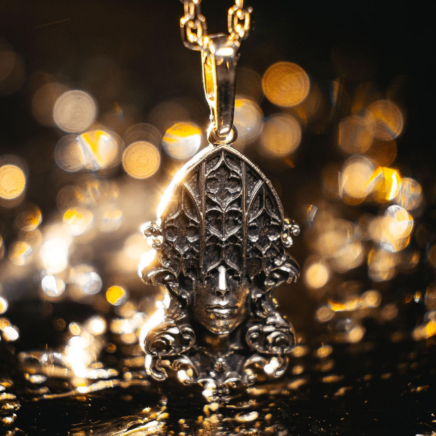 A detailed studio shot of a sterling silver pendant resting on a dark, textured stone surface. The design features a realistic human face in the center, but the forehead and eyes are obscured by a grid of Gothic window tracery (vertical bars and trefoils). The chin is framed by swirling Art Nouveau-style acanthus leaves. The silver is heavily oxidized, giving the face the appearance of a weathered stone statue. (etsy image id: 7448678222)