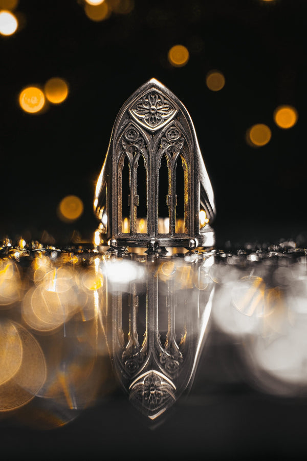 A clean studio shot of a tall, architectural sterling silver ring standing on a matte grey surface. The ring creates the silhouette of a Gothic "Lancet Window." It features three vertical openings separated by slender bars (mullions), topped by an intricate geometric tracery design within the pointed arch. The metal is matte silver with dark oxidation in the corners. (etsy image id: 7500328125)