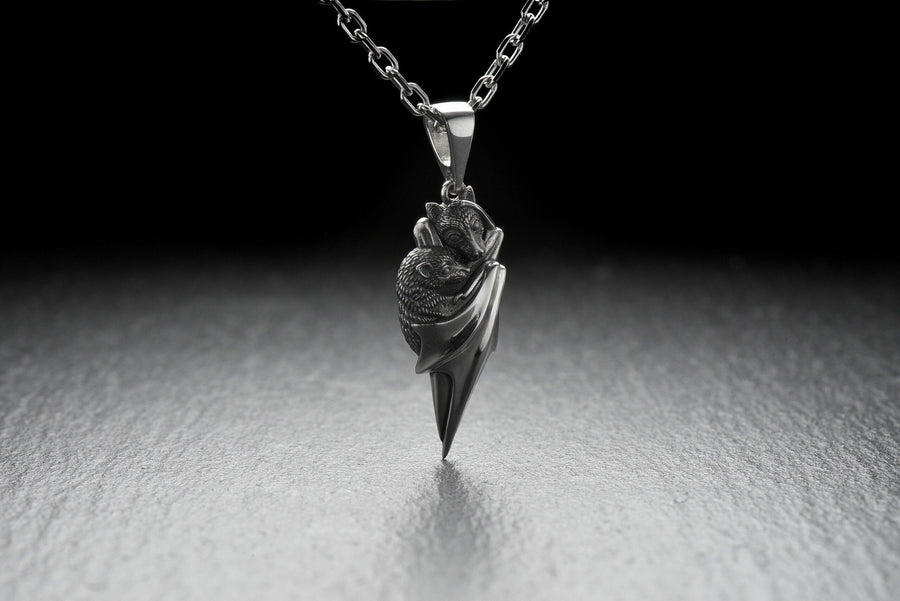 A detailed close-up studio shot of a sterling silver pendant shaped like two flying foxes or fruit bats. The bats are intimately huddled together, wrapped within the smooth, sweeping folds of their leathery wings. The metal is highly textured to show the fine details of their fur and faces. The silver is heavily oxidized in the recesses, creating a striking contrast against the polished highlights of the wings. (etsy image id: 5592925539)