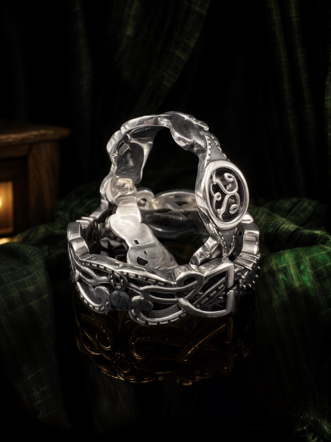 A studio shot of a matching two-piece sterling silver ring set on a black reflective surface. The standing ring is a signet style featuring an oval face with a raised Triskelion (triple spiral) motif. The ring lying flat is a wide band with an "openwork" or cut-out Celtic knot pattern. The silver is highly polished on the raised surfaces, contrasting with the dark shadows of the open areas.