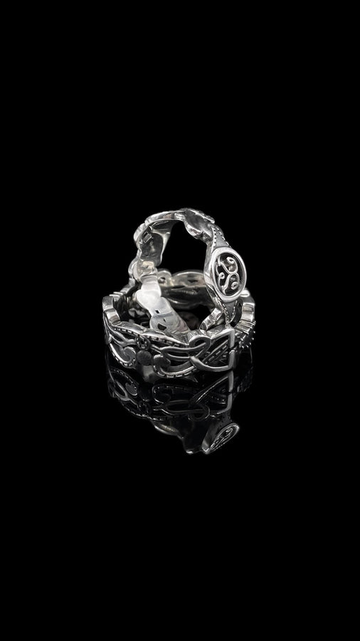 A studio shot of a matching two-piece sterling silver ring set on a black reflective surface. The standing ring is a signet style featuring an oval face with a raised Triskelion (triple spiral) motif. The ring lying flat is a wide band with an "openwork" or cut-out Celtic knot pattern. The silver is highly polished on the raised surfaces, contrasting with the dark shadows of the open areas.