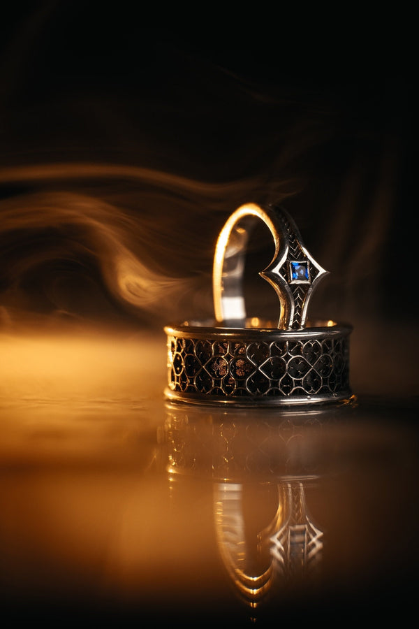 A high-definition shot of a matching two-piece wedding ring set resting on dark velvet. The foreground features a delicate sterling silver ring with a pointed, kite-shaped face holding a square blue gemstone. Standing behind it is a wide, sturdy band with a repeating quatrefoil "clover" pattern. Both rings share a cohesive finish: bright polished silver highlights against a deeply oxidized, matte black background.