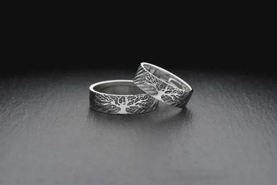 A clean studio shot of two matching sterling silver rings resting on a slate grey surface. Both bands feature a continuous panoramic design of a tree with spreading branches and deep roots. The tree motif is polished silver, which contrasts sharply with the textured, oxidized grey background that resembles tree bark or drifting soil.