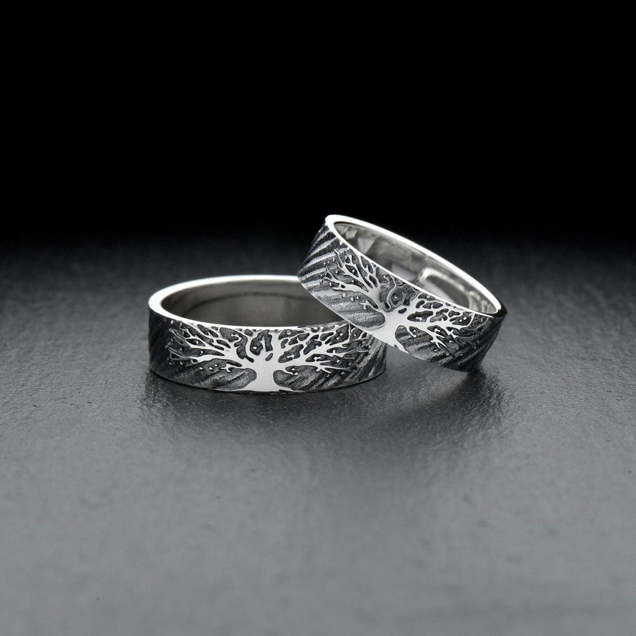 A clean studio shot of two matching sterling silver rings resting on a slate grey surface. Both bands feature a continuous panoramic design of a tree with spreading branches and deep roots. The tree motif is polished silver, which contrasts sharply with the textured, oxidized grey background that resembles tree bark or drifting soil.