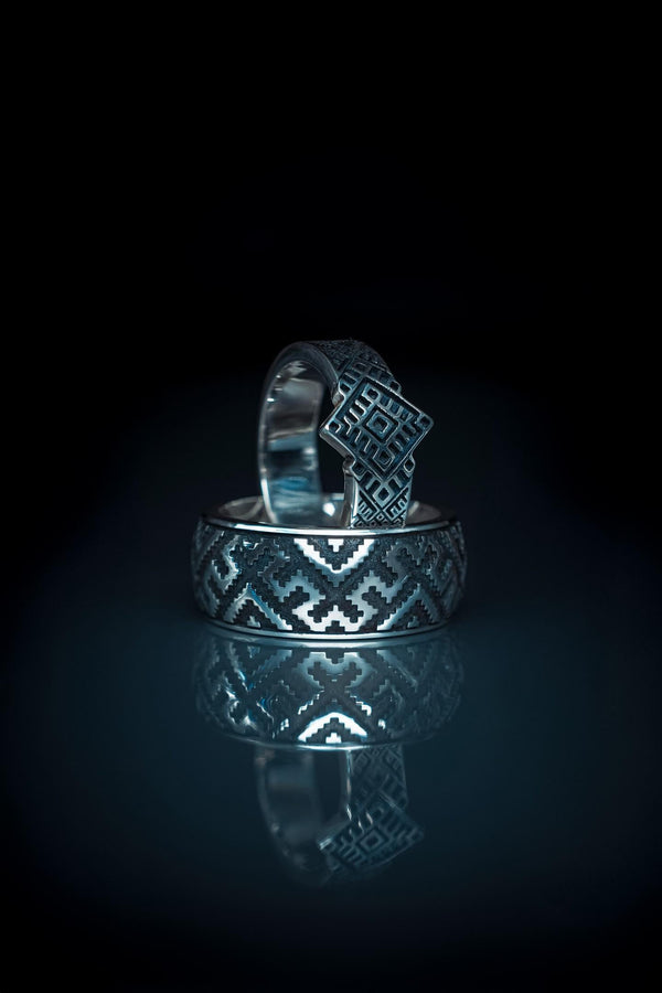 A studio shot of a two-piece sterling silver ring set with a high-contrast black and silver finish. The standing ring features a square, diamond-shaped face created by stepped geometric lines. The lying ring is a wide band with a continuous "zig-zag" or "step" pattern running around the circumference. The design mimics the look of cross-stitch embroidery or pixelated ancient art rendered in metal.