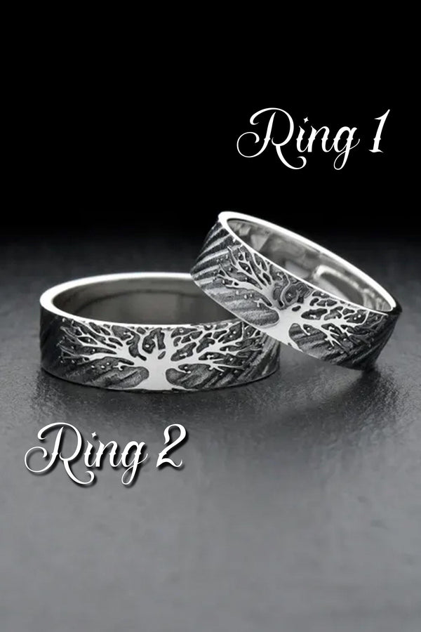 A clean studio shot of two matching sterling silver rings resting on a slate grey surface. Both bands feature a continuous panoramic design of a tree with spreading branches and deep roots. The tree motif is polished silver, which contrasts sharply with the textured, oxidized grey background that resembles tree bark or drifting soil.