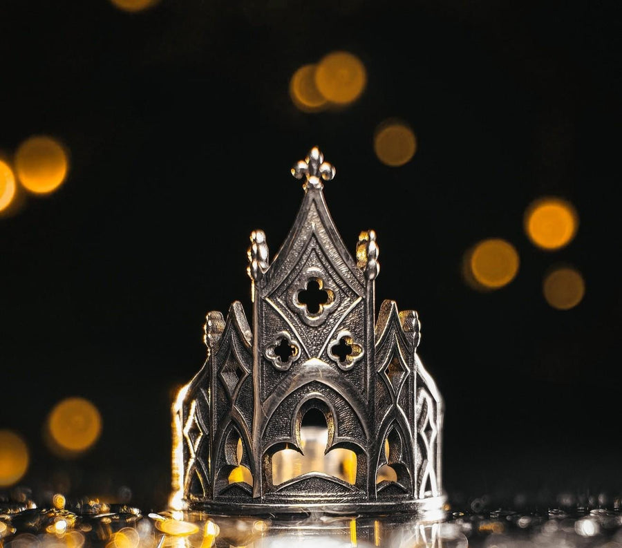 A clean studio shot of a sculptural sterling silver ring resting on a matte grey surface. The ring depicts a Gothic building facade with a tall, triangular central peak (gable) filled with geometric cross-shaped tracery. Flanking the center are lower pointed arches and vertical buttresses that wrap around the band. The silver is matte-finished with dark oxidation in the recesses to mimic stone shadows. (etsy image id: 7452370200)