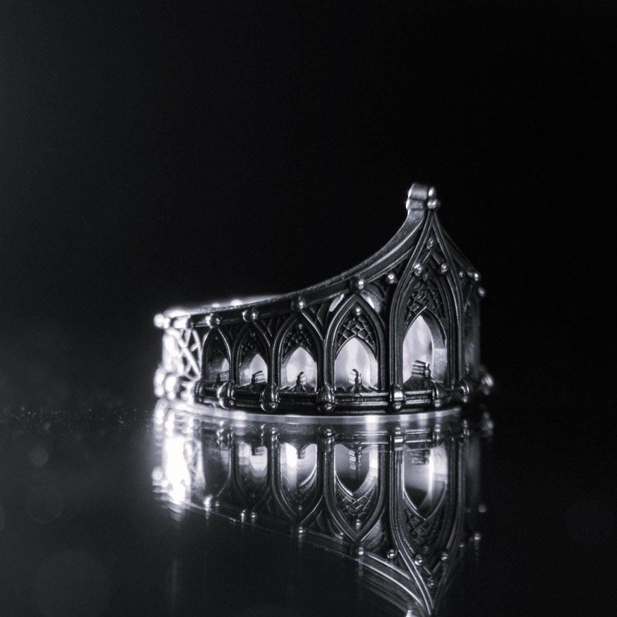 A cinematic studio shot of a sculptural sterling silver ring standing on a reflective surface. The ring is designed as a "crown" or "coronet," formed by a continuous circle of Gothic cathedral arches. The central arch is the tallest, creating a focal point like a spire. The silver is heavily oxidized, with black recesses emphasizing the ribbed vaulting and architectural details against the polished ridges.