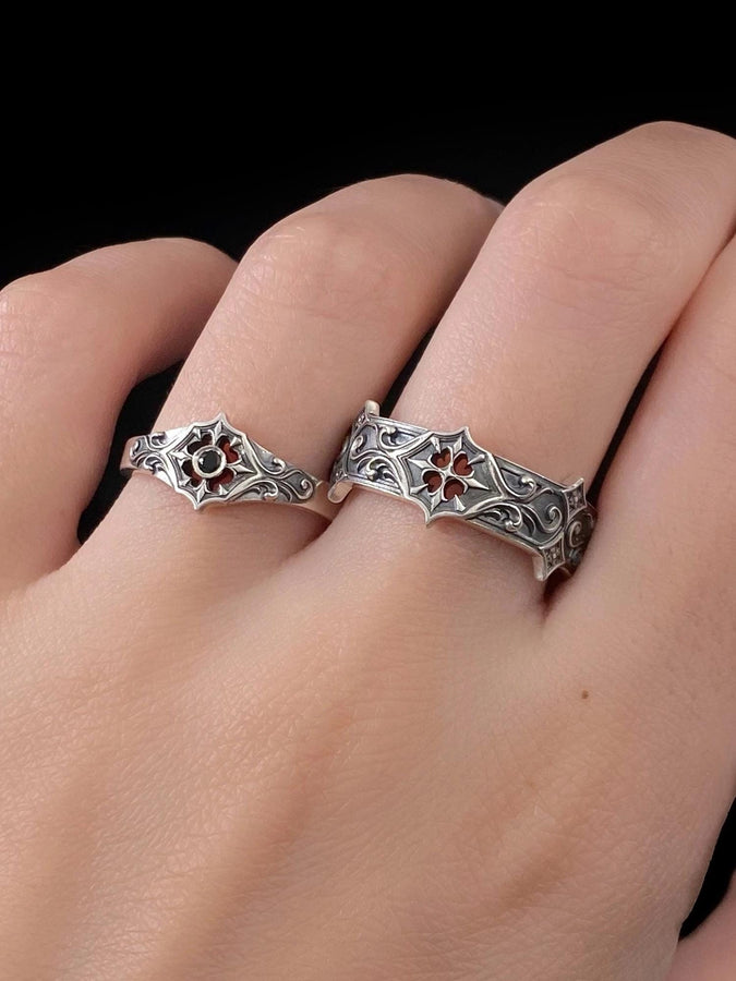 A cinematic studio shot of a matching two-piece ring set on dark velvet. The top ring stands upright, featuring a diamond-shaped face with a four-pointed star design and a small, round black stone in the center. The bottom ring lies flat, displaying a wide band with a repeating pattern of the same star/cross motif connected by swirling filigree. The silver is polished on the ridges and dark in the background.