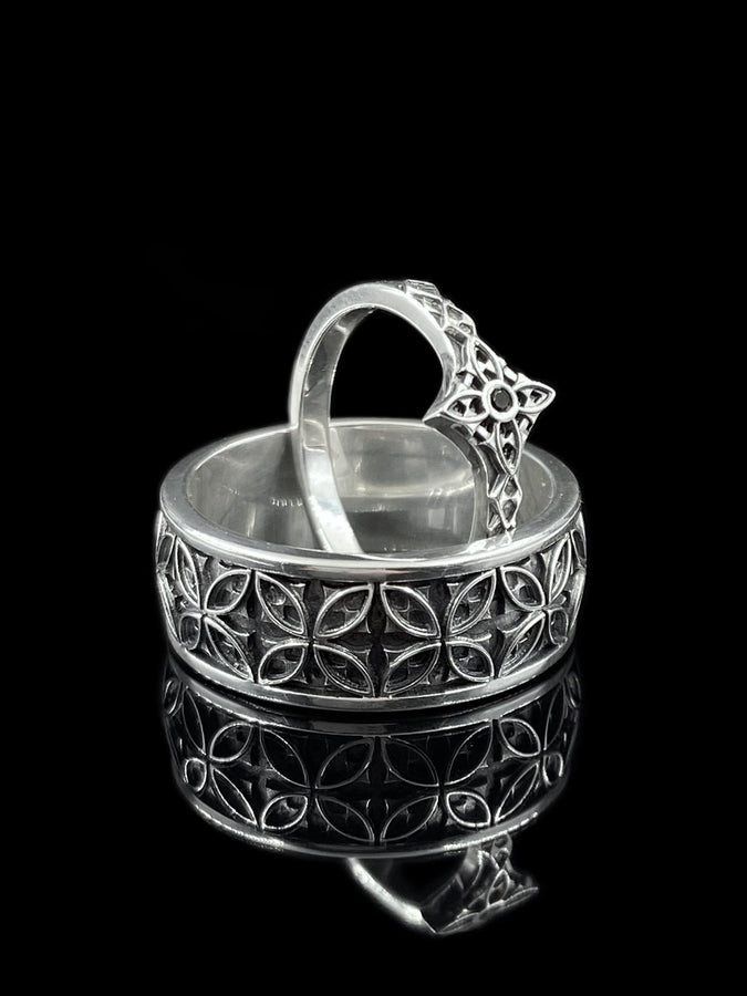 A professional studio photograph of a matching two-piece sterling silver wedding ring set. Both rings feature a prominent, raised Greek-style cross motif in the center, which holds a round, bezel-set white crystal. The interior of the cross arms has a fine, cross-hatched grid texture. Along the sides of the high-polished silver bands are recessed, four-lobed quatrefoil shapes. The rings are presented against a pitch-black background, making the p __etsy:8051335185