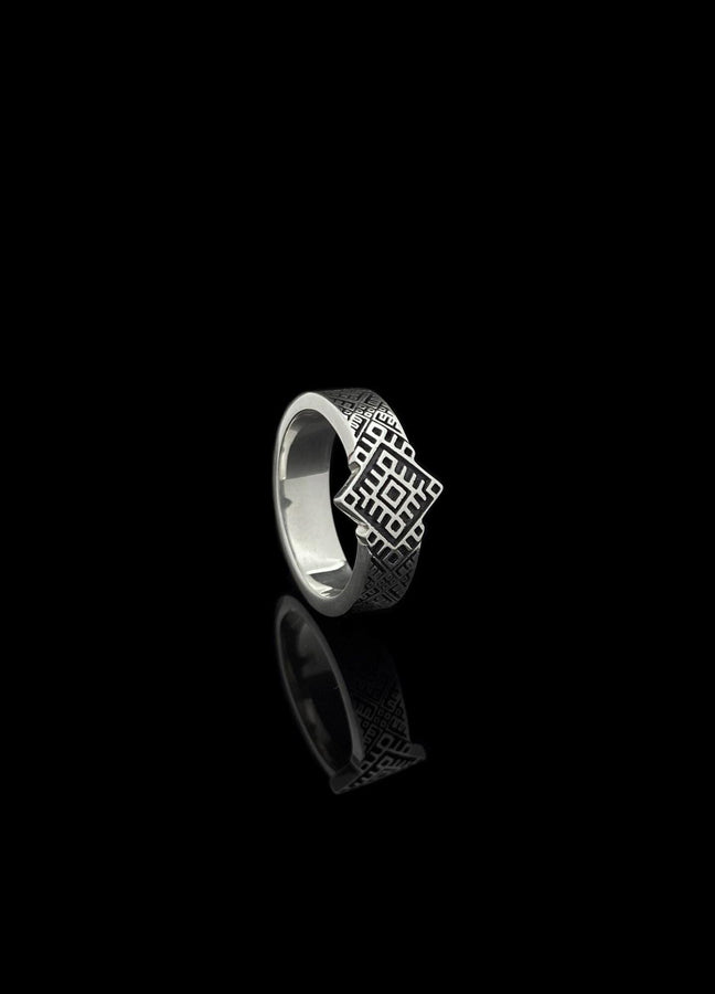 A studio shot of a two-piece sterling silver ring set with a high-contrast black and silver finish. The standing ring features a square, diamond-shaped face created by stepped geometric lines. The lying ring is a wide band with a continuous "zig-zag" or "step" pattern running around the circumference. The design mimics the look of cross-stitch embroidery or pixelated ancient art rendered in metal.